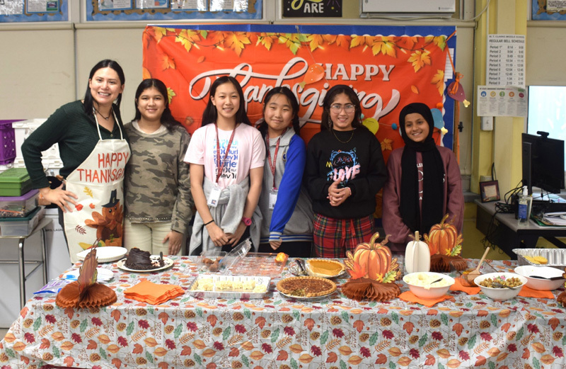 Group of six people smiling in front of a festive "Happy Thanksgiving" banner. A table with autumn-themed decor and various dishes is in the foreground.