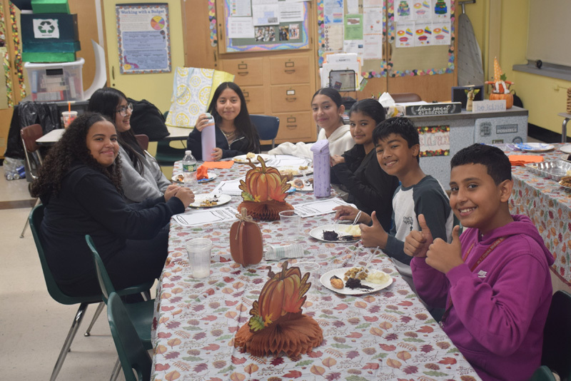 Seven kids sit around a festive table with autumn-themed decorations and paper pumpkins. They smile and show thumbs-up, conveying a joyful atmosphere.