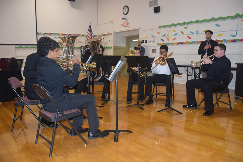 A large group of smiling students poses indoors, holding certificates. They wear matching "champions" shirts. A colorful mural is on the right wall.