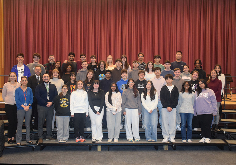 A diverse group of teenagers and adults stand on risers against a purple curtain backdrop, smiling warmly, conveying a sense of camaraderie and community.