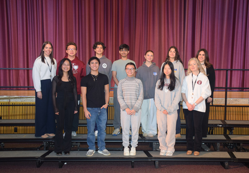A diverse group of teenagers and adults stand on risers against a purple curtain backdrop, smiling warmly, conveying a sense of camaraderie and community.