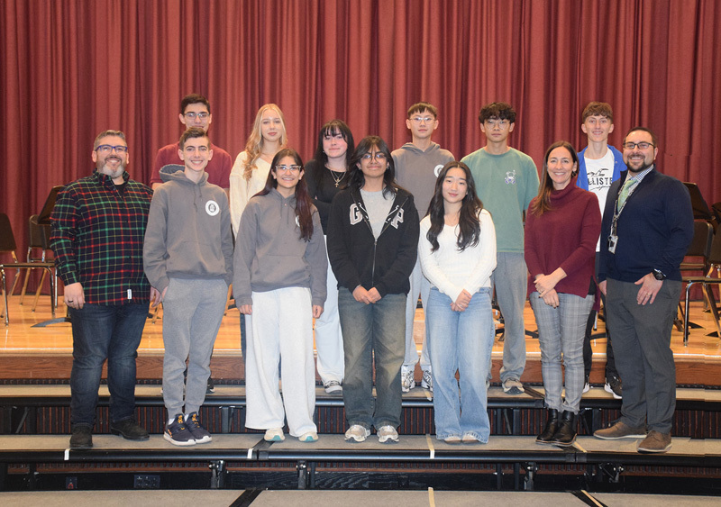 A diverse group of teenagers and adults stand on risers against a purple curtain backdrop, smiling warmly, conveying a sense of camaraderie and community.
