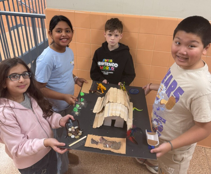 children proudly display a detailed diorama featuring a rustic wooden house, trees, and tiny figures. 
