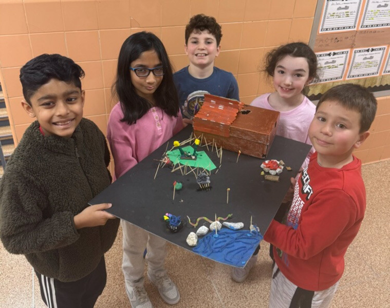 children proudly display a detailed diorama featuring a rustic wooden house, trees, and tiny figures. 