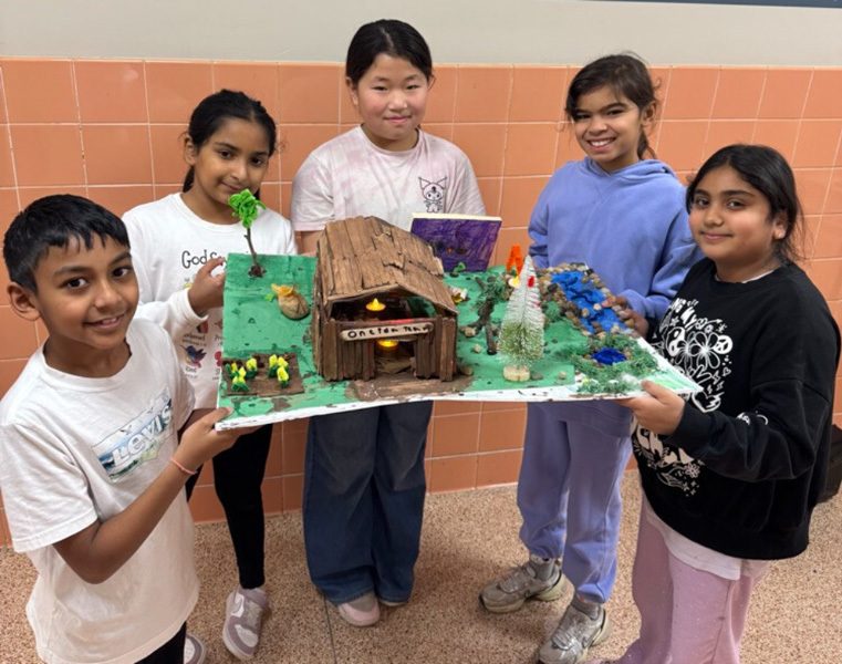 children proudly display a detailed diorama featuring a rustic wooden house, trees, and tiny figures. 