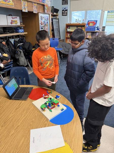 A bustling classroom filled with students and a teacher engaging in various activities. Some students use laptops, while others chat or examine projects, creating an energetic and collaborative atmosphere.
