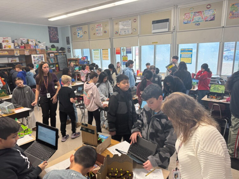 A bustling classroom filled with students and a teacher engaging in various activities. Some students use laptops, while others chat or examine projects, creating an energetic and collaborative atmosphere.
