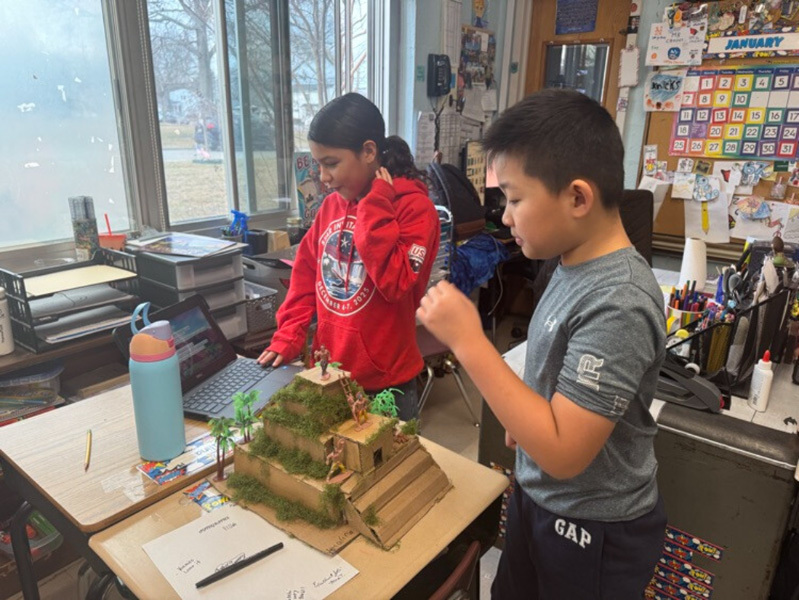 A bustling classroom filled with students and a teacher engaging in various activities. Some students use laptops, while others chat or examine projects, creating an energetic and collaborative atmosphere.