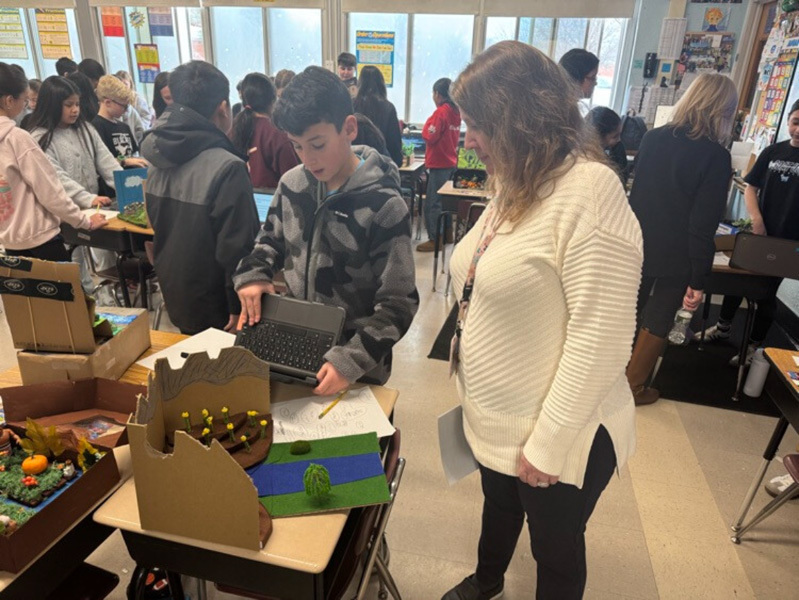 A bustling classroom filled with students and a teacher engaging in various activities. Some students use laptops, while others chat or examine projects, creating an energetic and collaborative atmosphere.