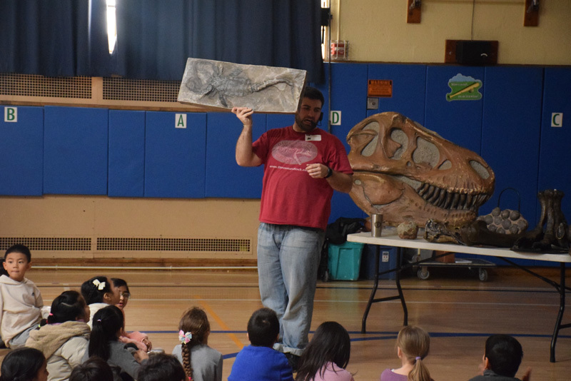 Fossils and dinosaur replicas are displayed on a table, with a projection screen showing related images in the background, suggesting an educational setting focused on paleontology.