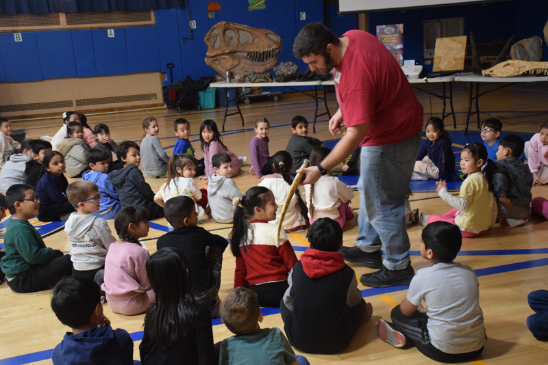 Fossils and dinosaur replicas are displayed on a table, with a projection screen showing related images in the background, suggesting an educational setting focused on paleontology.