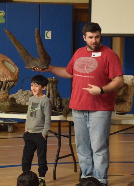 Fossils and dinosaur replicas are displayed on a table, with a projection screen showing related images in the background, suggesting an educational setting focused on paleontology.