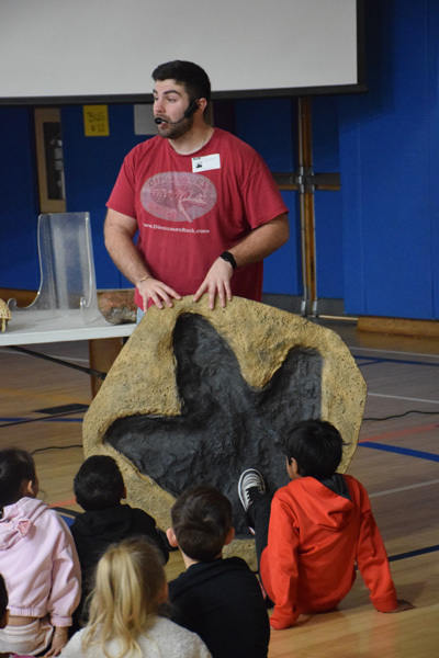 Fossils and dinosaur replicas are displayed on a table, with a projection screen showing related images in the background, suggesting an educational setting focused on paleontology.