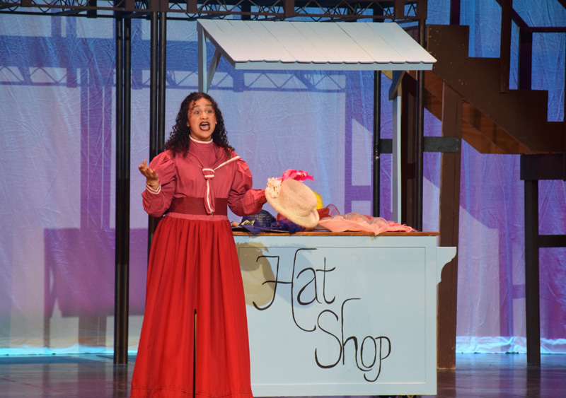 Theatrical performance with actors in colorful, vintage costumes and hats, standing in front of a red curtain. 