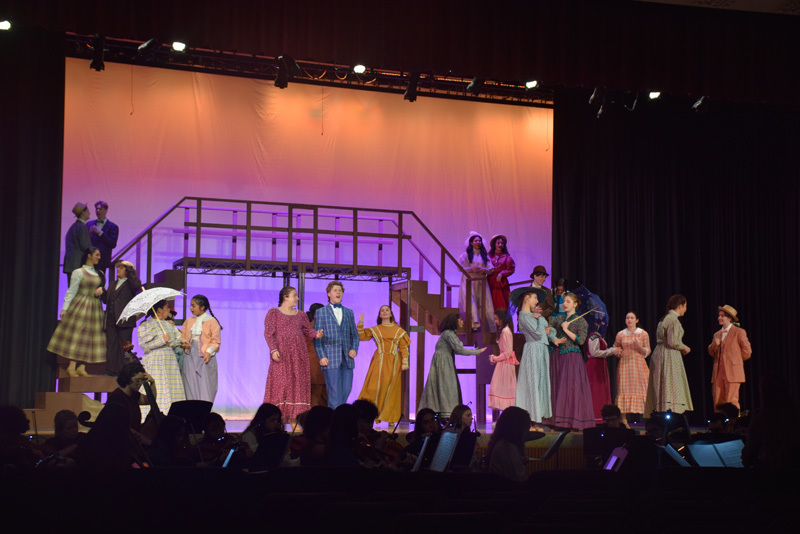 Theatrical performance with actors in colorful, vintage costumes and hats, standing in front of a red curtain. 
