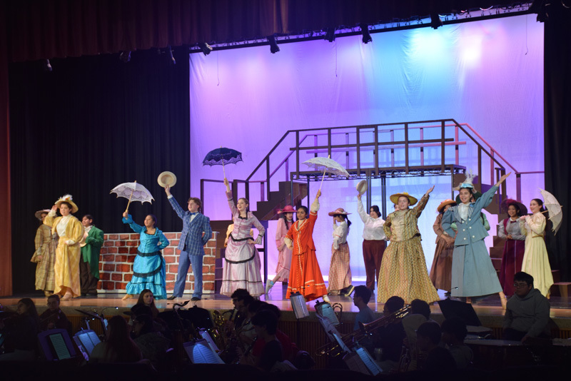Theatrical performance with actors in colorful, vintage costumes and hats, standing in front of a red curtain. 