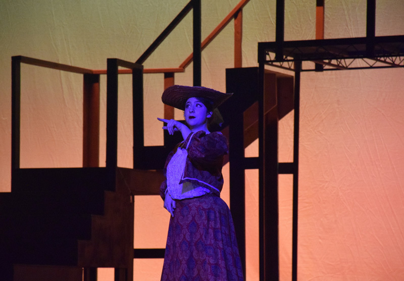 Theatrical performance with actors in colorful, vintage costumes and hats, standing in front of a red curtain. 