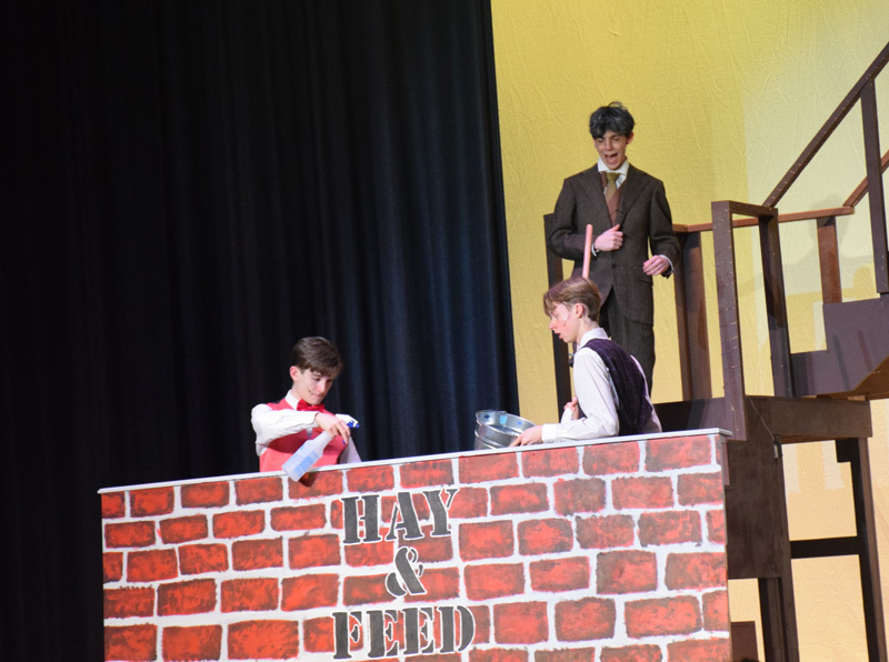Theatrical performance with actors in colorful, vintage costumes and hats, standing in front of a red curtain. 