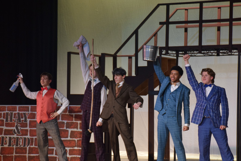 Theatrical performance with actors in colorful, vintage costumes and hats, standing in front of a red curtain. 