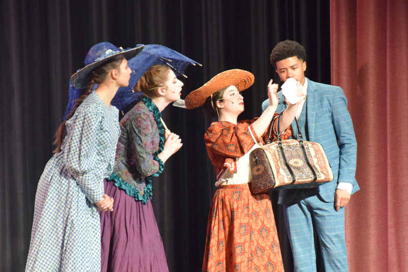 Theatrical performance with actors in colorful, vintage costumes and hats, standing in front of a red curtain. 