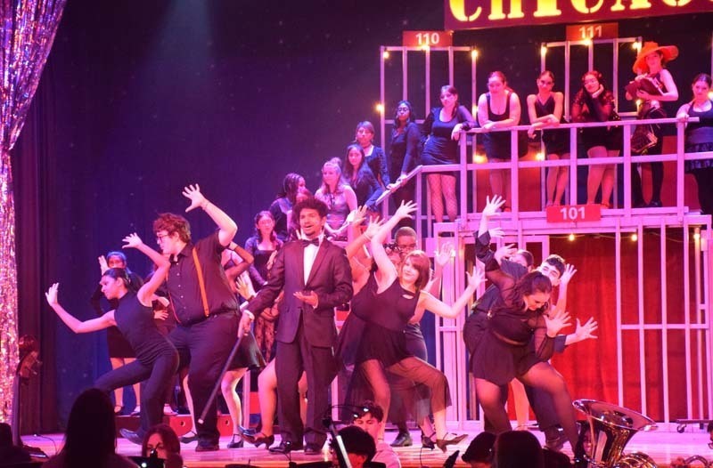 Photo of students wearing suits, dresses and red and black clothing performing a musical on stage.