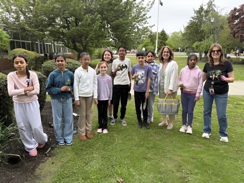 Group of children and adults participating in a community garden project, smiling and holding plants outdoors on a sunny day.