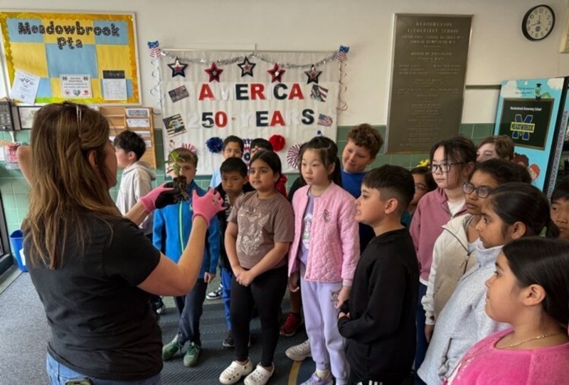 Elementary school students gather as a teacher demonstrates a lesson about plants in front of an "America 50 Years" banner.