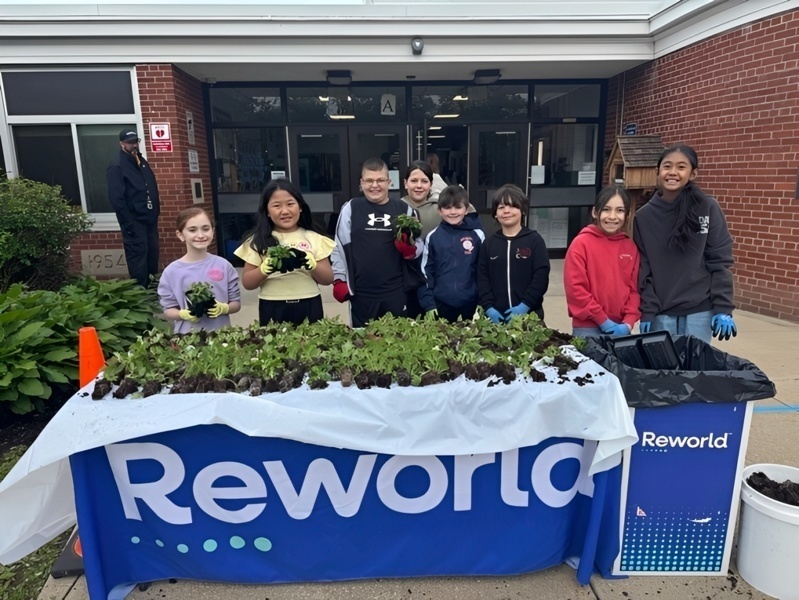 Students participate in a community planting event, showcasing their plants at a table with the "Reworld" banner outside a school building.