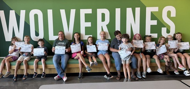 Kids sitting in front of a wolverine sign, smiling with their certificates