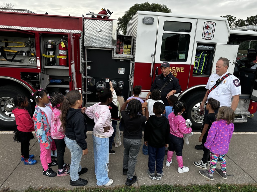 Kindergarten students surrounding and exploring a red fire truck with  firefighters standing nearby during a visit from the East Moline Fire Department.