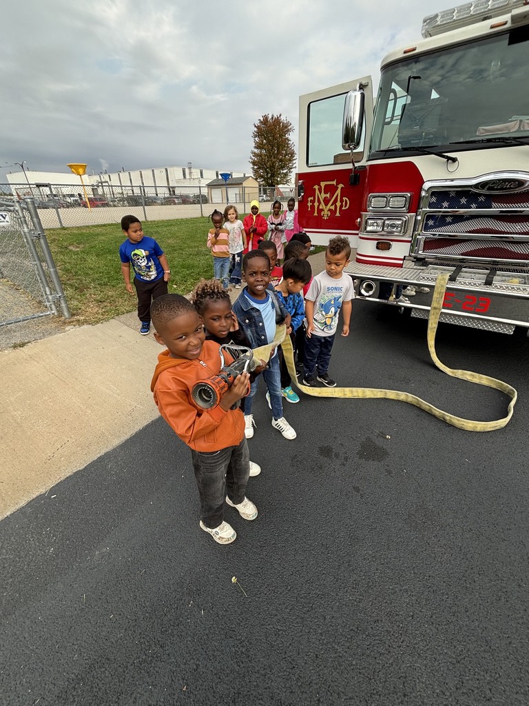 Kindergarten students surrounding and exploring a red fire truck with  firefighters standing nearby during a visit from the East Moline Fire Department.
