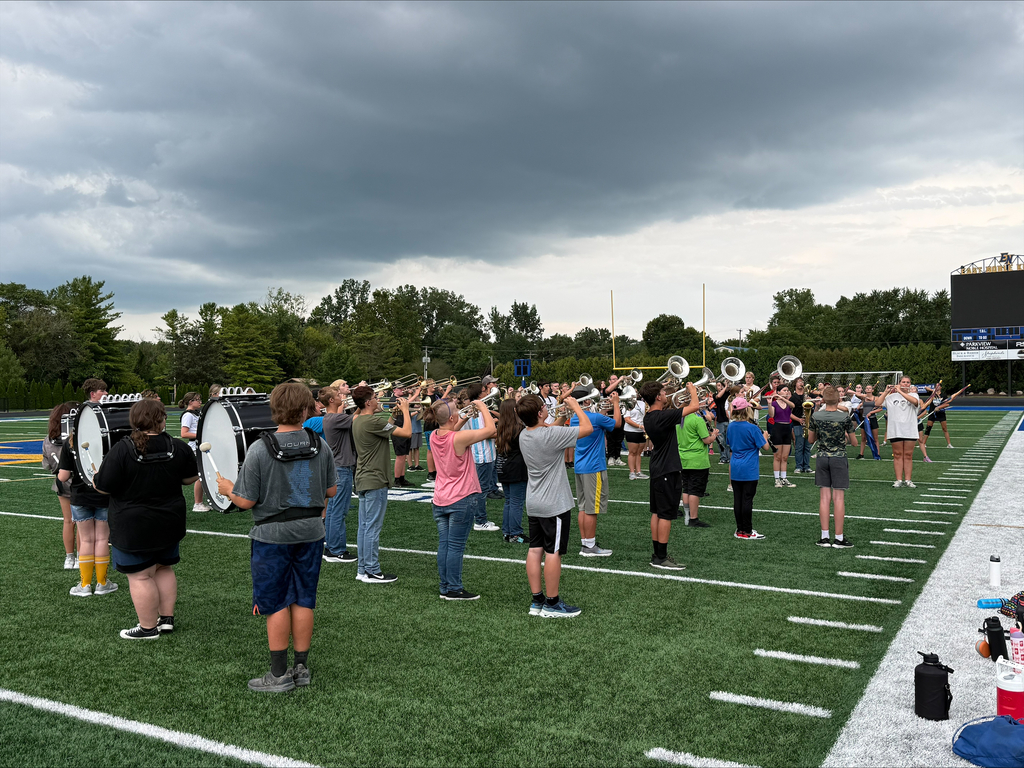 Marching band students playing their instruments while marching in a precise, synchronized formation on the football field.