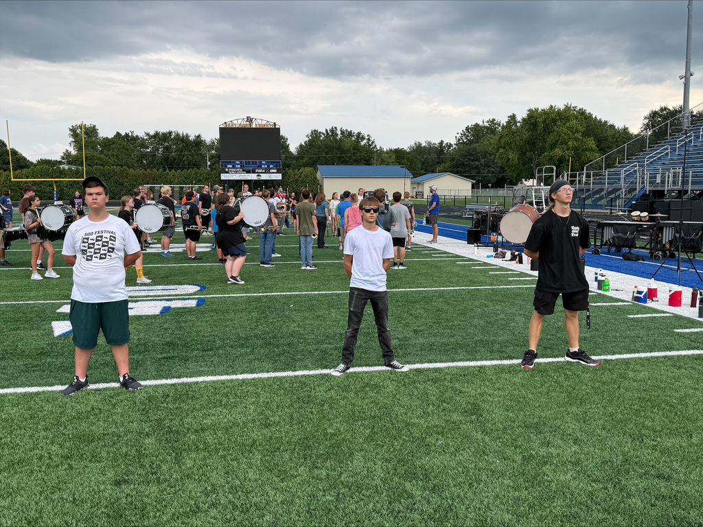 High school marching band students standing at parade rest with feet apart, hands behind their backs, and chins held high in a disciplined, proud posture.