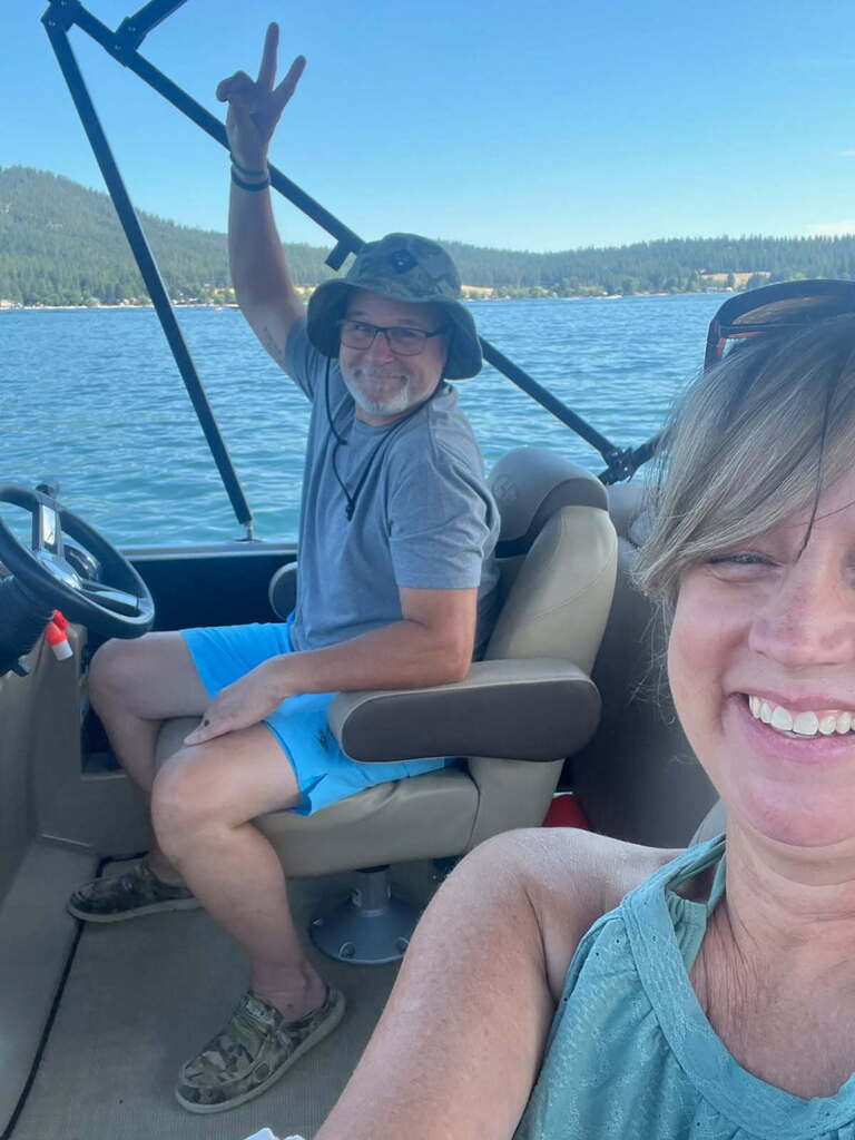 Two people on a boat, one wearing a hat, smiling and giving a peace sign. Behind them, mountains and water.