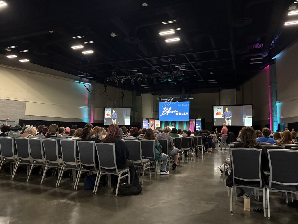 An audience seated in rows of chairs in a room, with screens on the wall showing presentations.