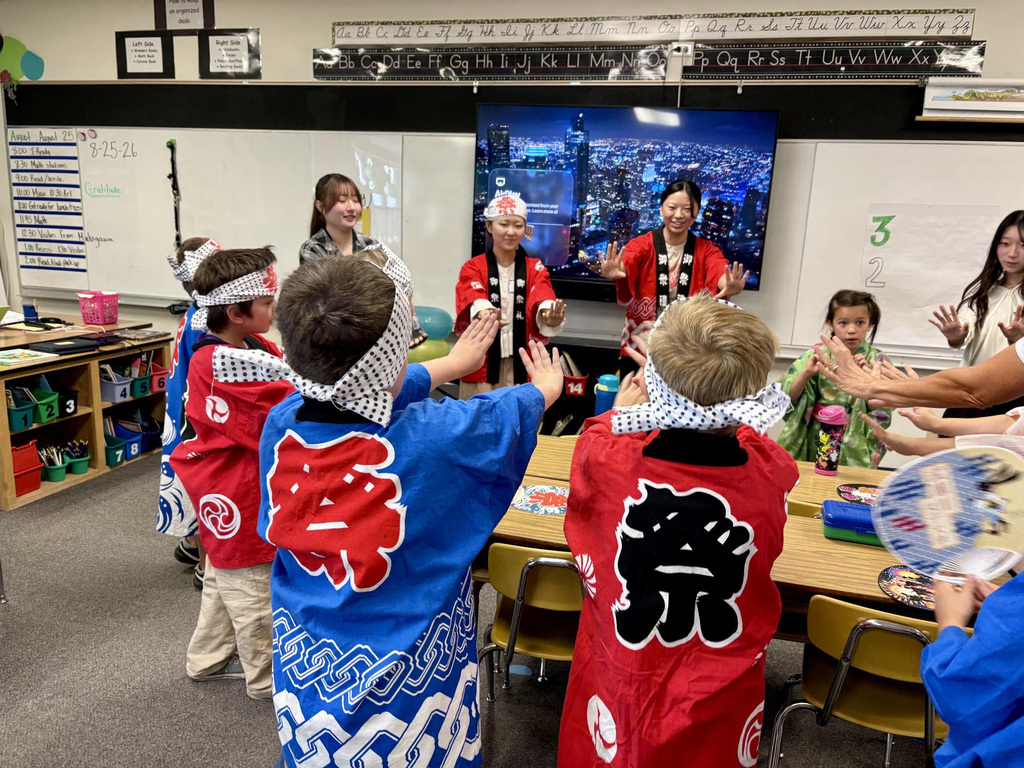 Children in traditional Japanese garb dance in a classroom, facing a screen displaying a city skyline.