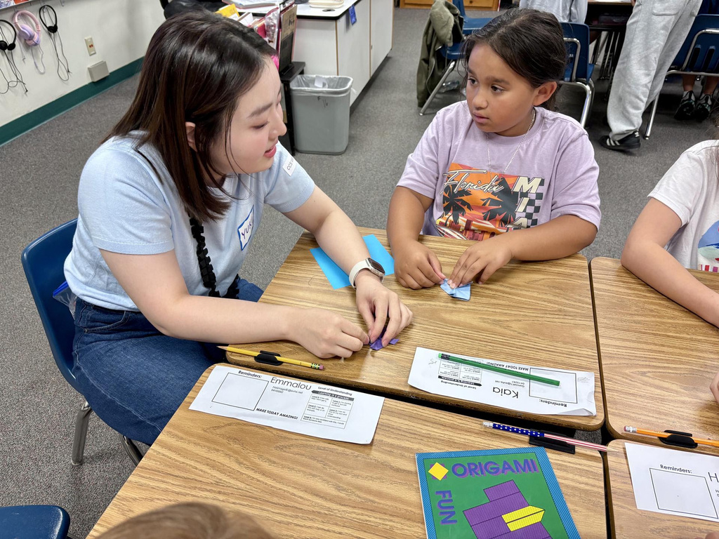A woman helps a child with a paper craft at a desk, surrounded by other students.