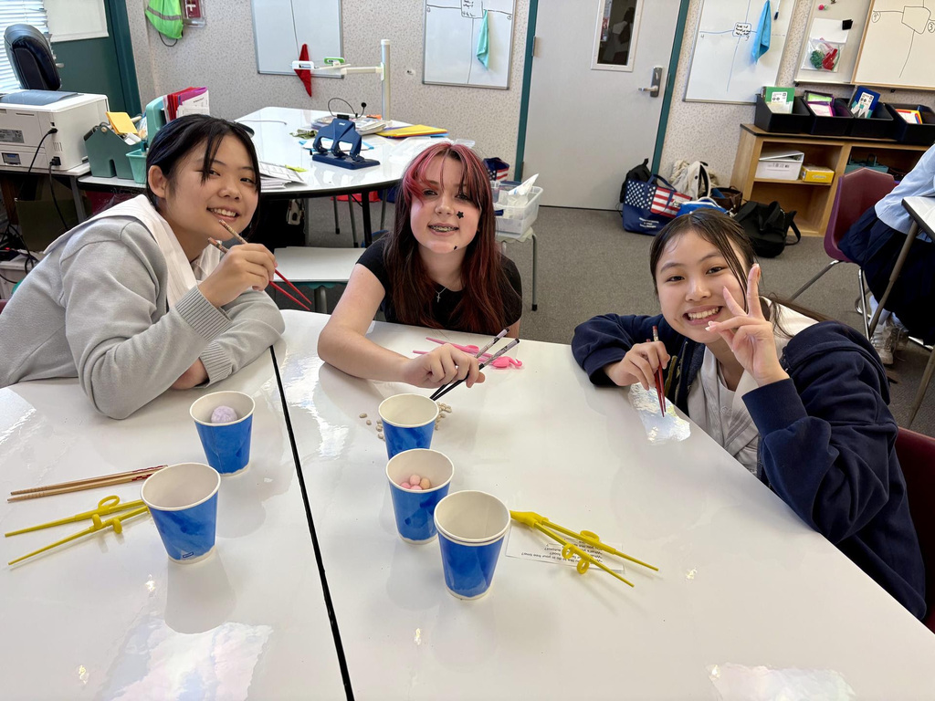Three students with paint on their faces seated at a table with cups, chopsticks, and paintbrushes.