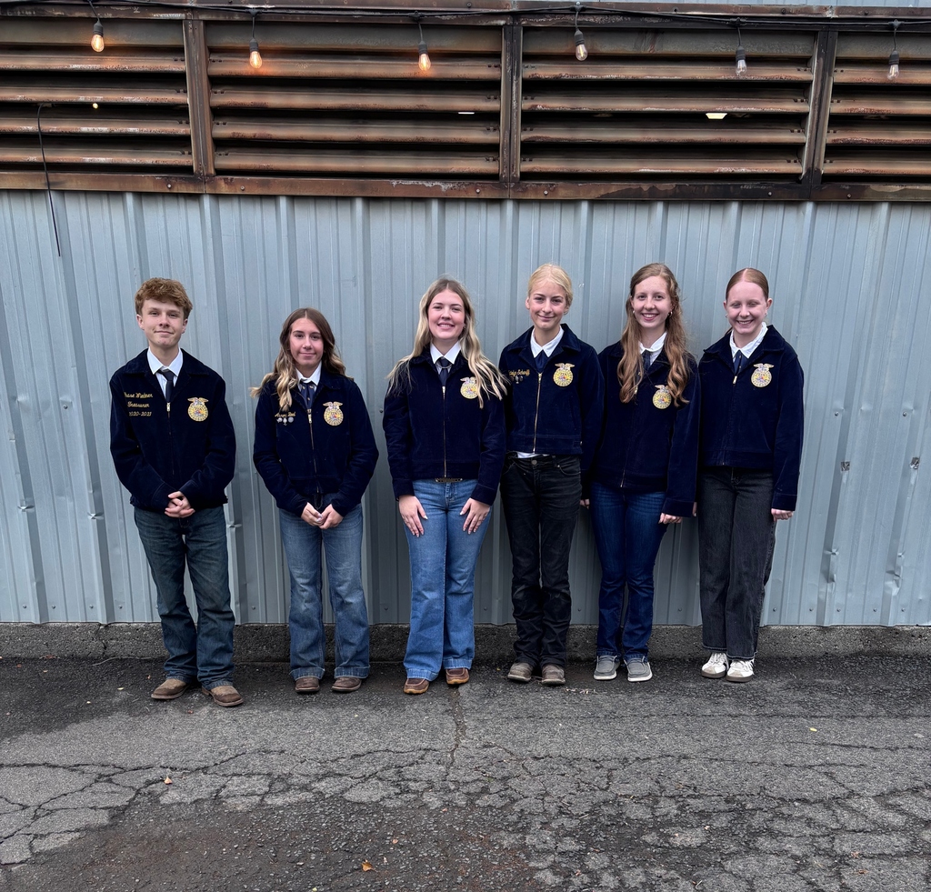 Six people in matching jackets and jeans pose against a silver wall with horizontal metal slats.