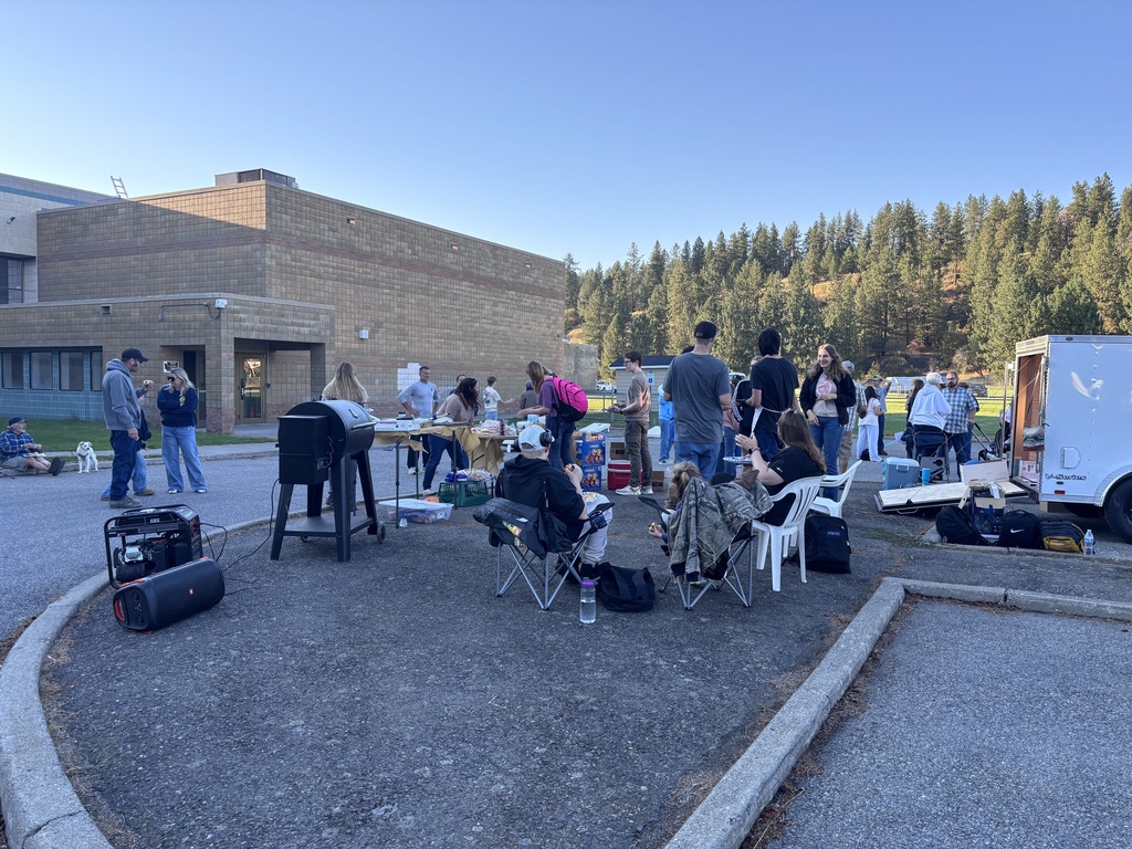 Group of people sitting and standing around tables in a parking lot. Building and trees in the background.