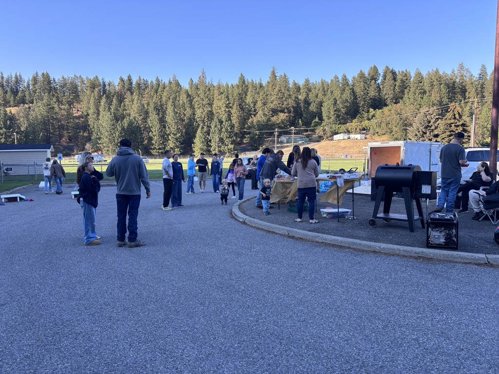 Many people are gathered in a parking lot with trees in the background. Some are standing near a grill.