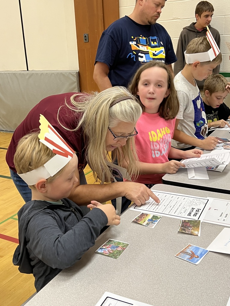 Group of kids wearing paper crowns, one adult helping, sitting around a table with cards and papers.