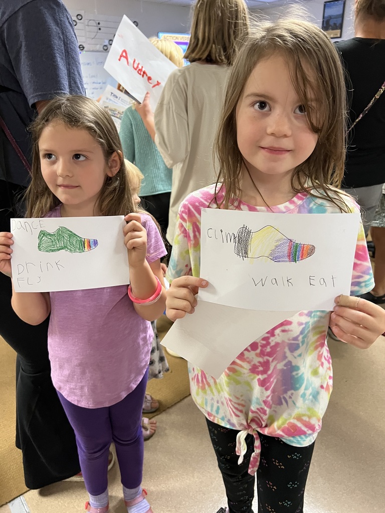 Two young girls show drawings of shoes with captions. Behind them are several people, some of whom are holding signs.