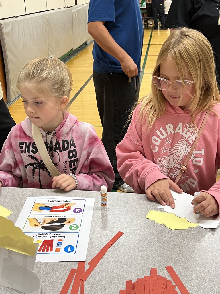 Two girls, one wearing glasses, seated at a table. They are using colored papers and glue.