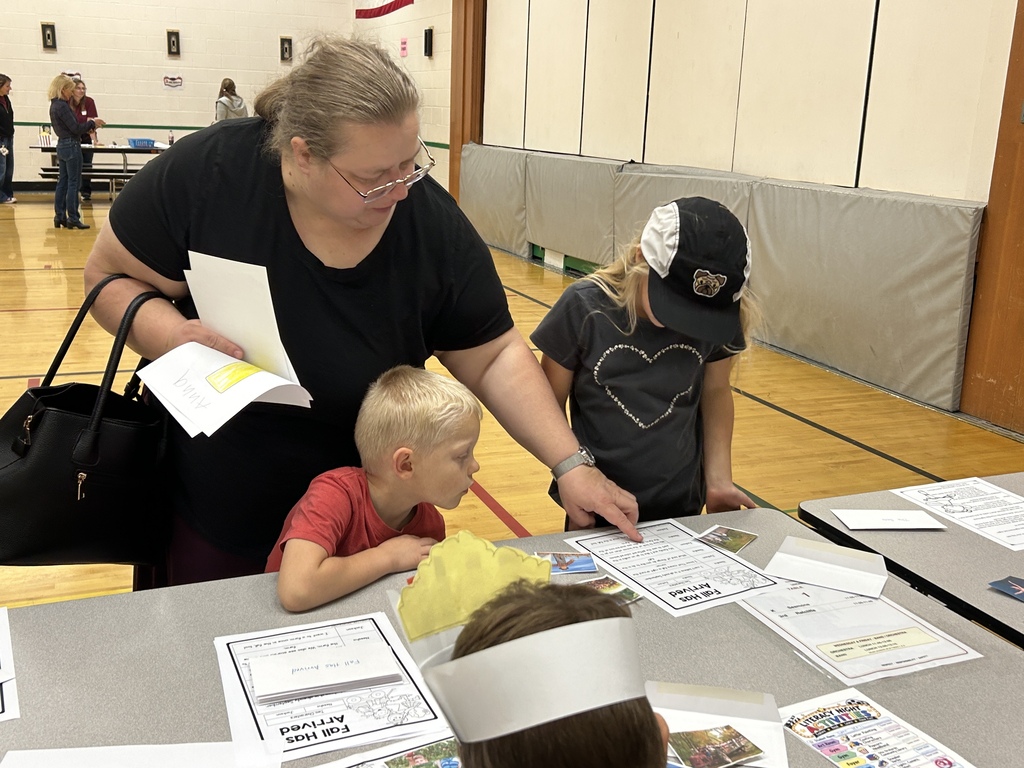 Woman wearing glasses and a black shirt helps two children with a project at a table.