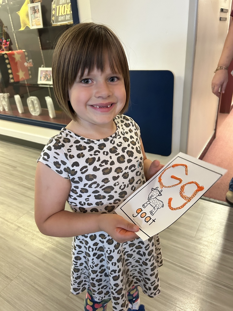 A young girl with short hair holds a card with the letters "gg" on it. She is smiling.