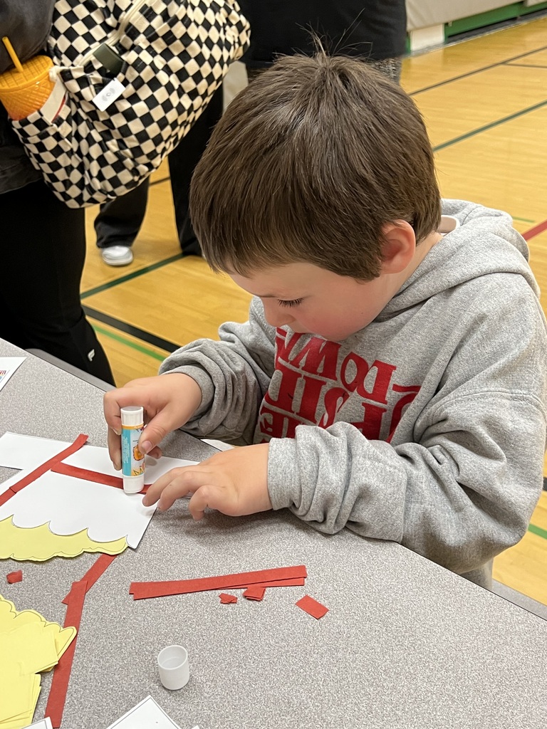 A young boy sits at a table, focused on a craft project. He holds a glue bottle and paper cutouts.