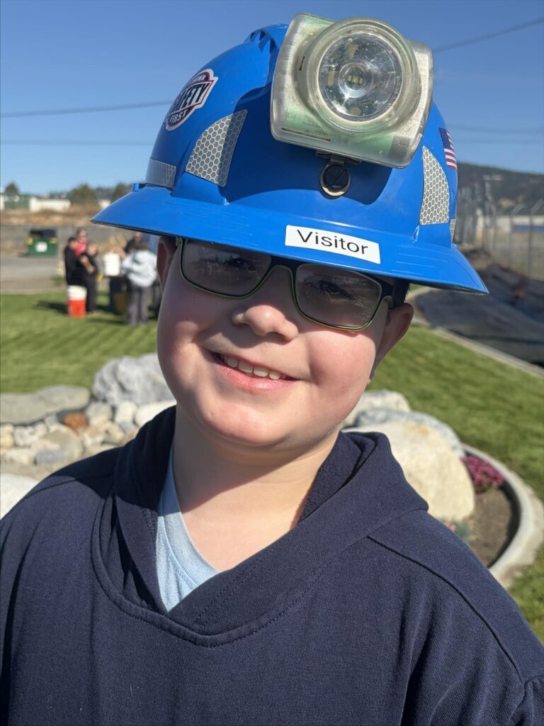Child wearing a blue hard hat with a lamp, smiling. Behind, a grassy area with rocks and people.