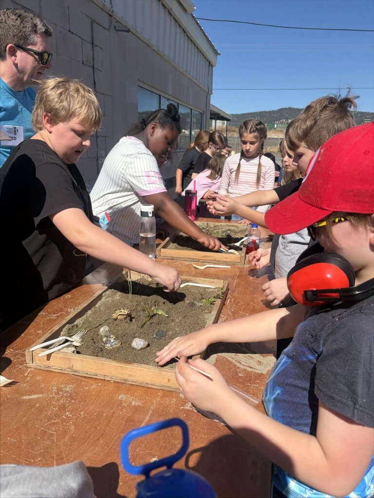 Children gather around a table outside, with some wearing hats, glasses, and ear protection, sifting through sand.