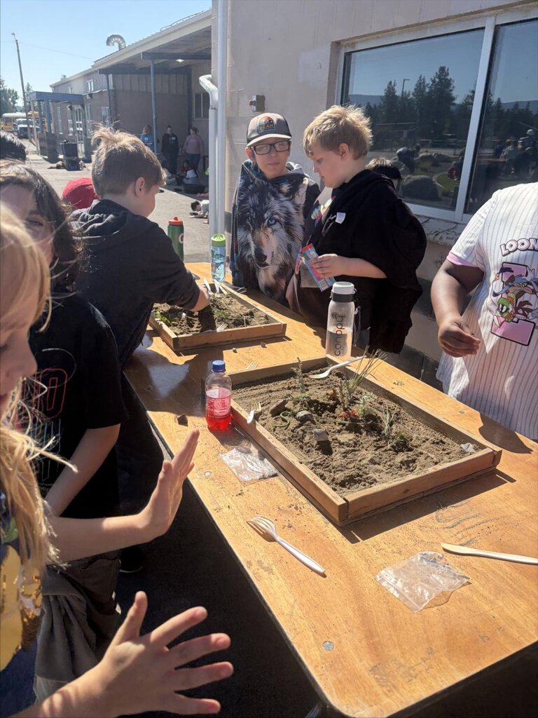 Kids gathered around a table with trays of soil and objects, engaging in an outdoor activity.